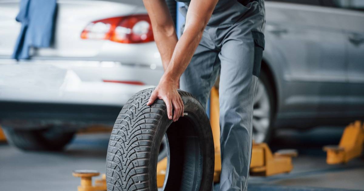 mecanic auto cu uniforma gri, invarte un cauciuc de masina. in spatele lui este o masina gri