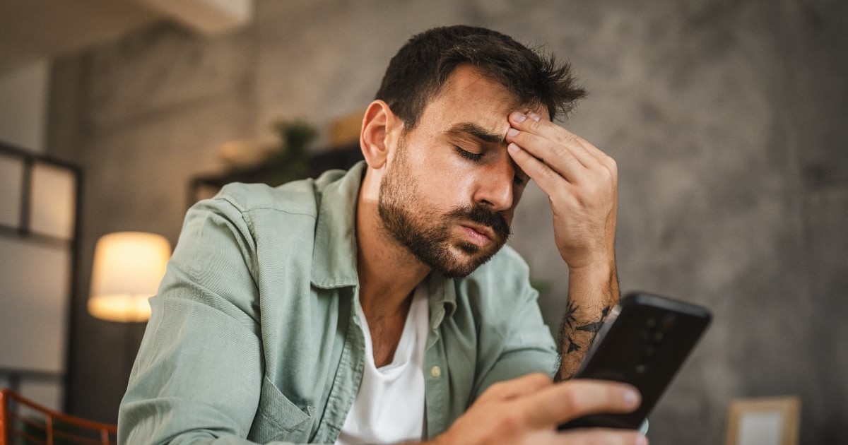 Un bărbat stă aplecat, ținându-și fruntea cu mâna în timp ce privește îngrijorat ecranul telefonului. Expresia sa sugerează stres și oboseală, ca și cum ar fi copleșit de informațiile primite.