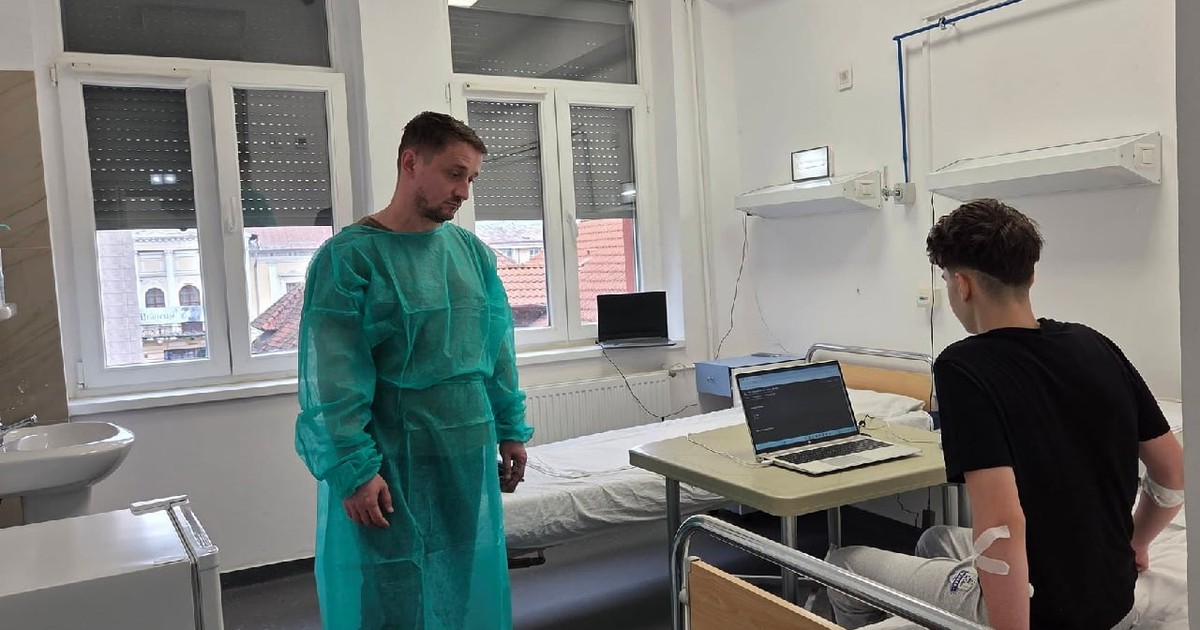 Un bărbat cu halat verde stă în fața unui băiat ămbracat cu tricou negru, care stă pe un pat de spital cu un laptop pe o măsuta din fata lui