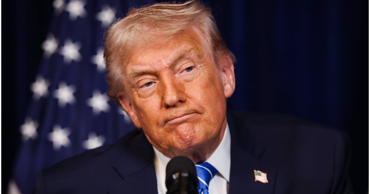 Portret în plan apropiat al lui Donald Trump având o expresie serioasă în fața unui microfon, cu steagul Statelor Unite ale Americii vizibil în fundalul albastru.