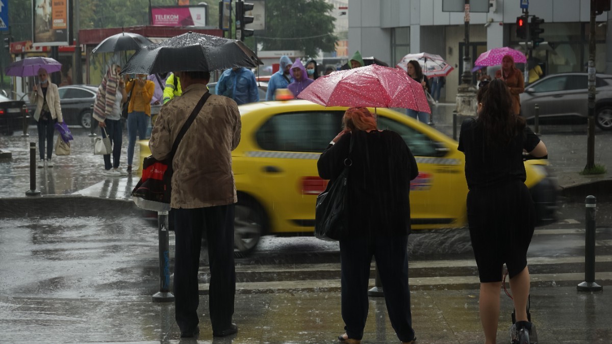 Pietoni îmbrăcați gros, cu umbrelele desfăcute, stau și așteaptă să traverseze la semafor, într-o zi ploioasă în București