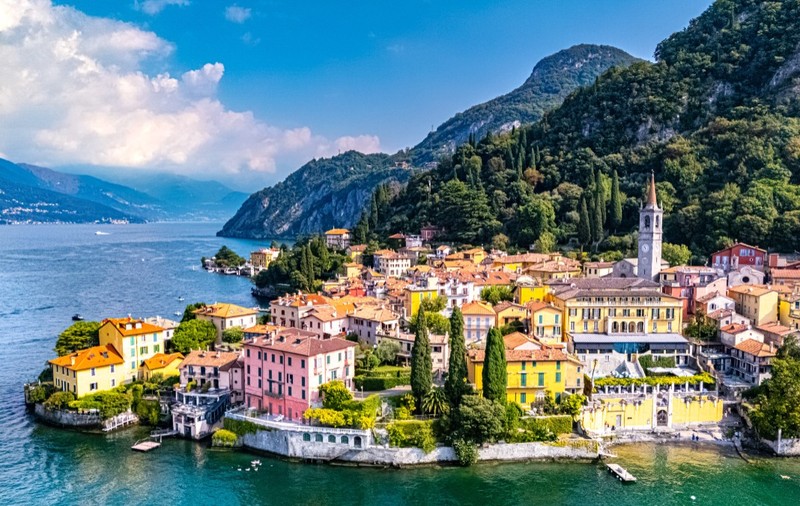 Imagine văzută de sus asupra orașului Varenna de pe malul Lacului Como din Italia, cu clădiri colorate și munți pe fundal.