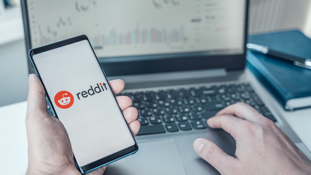 Ce este platforma Reddit şi cum să o foloseşti - Imagine cu un bărbat aflat în faţa unui laptop şi care ţine un telefon ce afişează logo-ul reddit