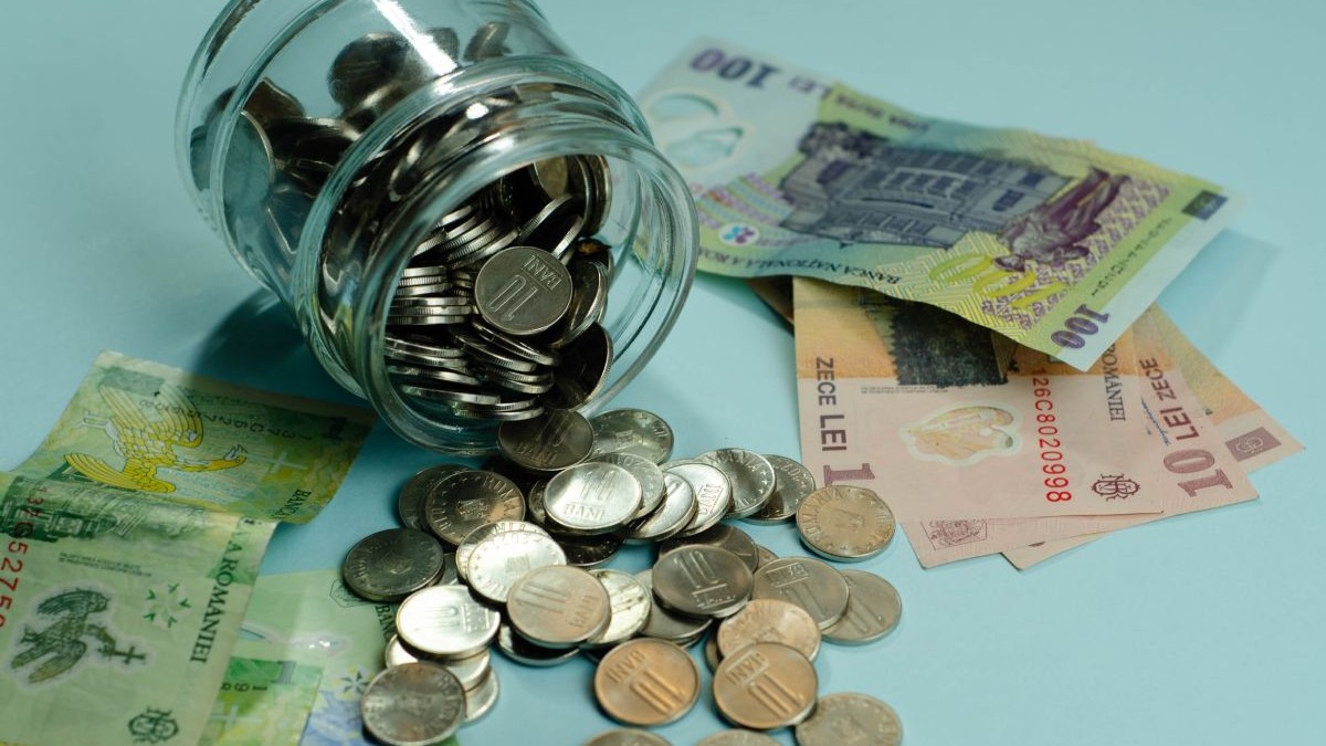 Borcan de sticlă cu monede și bancnote de 1 leu, 10 lei și o bancnotă de 100 de lei, pe o masă albastră