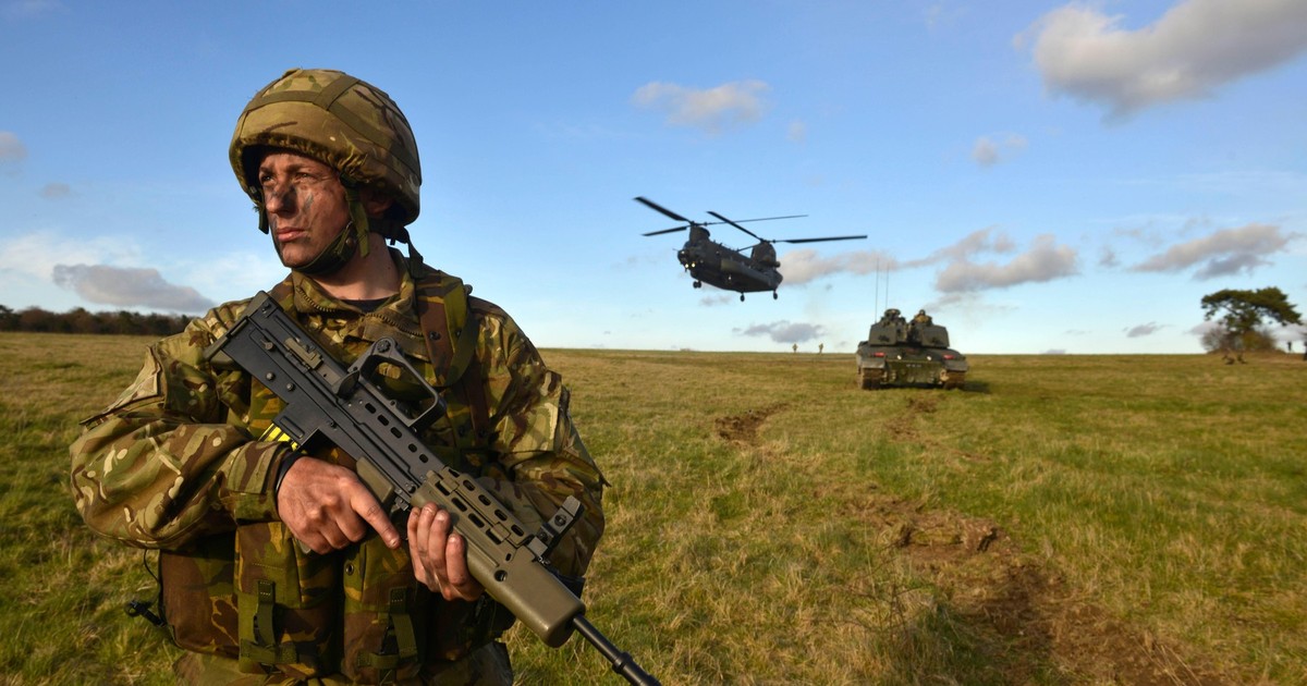 Soldat britanic surprins în timp ce ține în mâini o armă, iar în spatele său se află un elicopter și un tanc