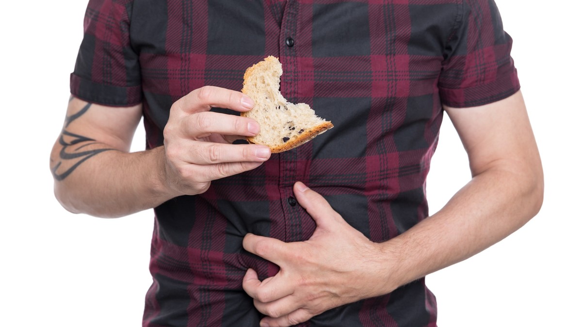 Alimente fără gluten – ce poți mânca dacă ai intoleranță la gluten - Imagine cu un bărbat care ţine într-o mână o felie muşcată de pâine şi cealaltă mână se ţine de burtă, sugerând intoleranţa la gluten