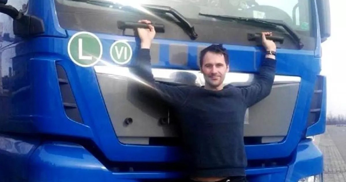 Un bărbat se ține cu mâinile de cabina unui camion albastru, stând în picioare în fața ei