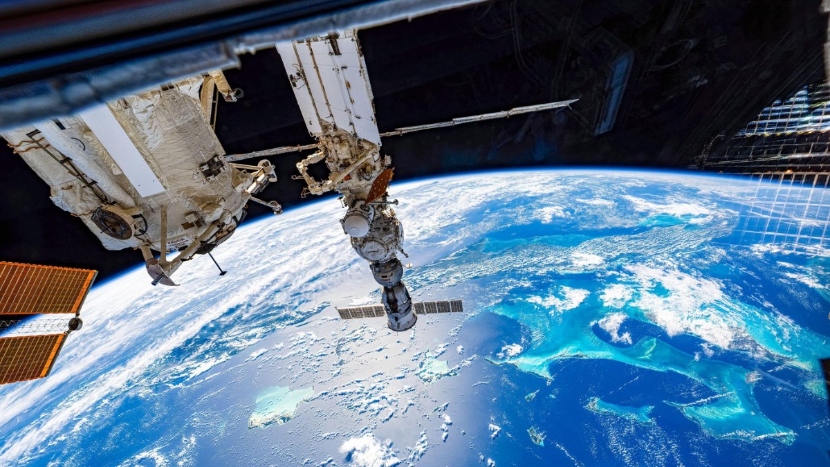 Imaginea surprinde Stația Spațială Internațională pe orbită, cu modulele și panourile sale solare vizibile deasupra Pământului. În fundal se vede planeta albastră, cu oceane și nori, oferind o perspectivă spectaculoasă asupra atmosferei și spațiului cosmic.