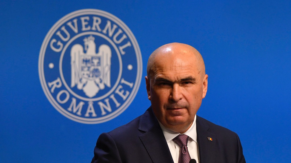 Ilie Bolojan surprins în fața unui microfon în timpul unei conferințe de presă la Palatul Victoria, în iulie 2025. Poartă un sacou negru, cu o cămașă albă și o cravată mov cu buline