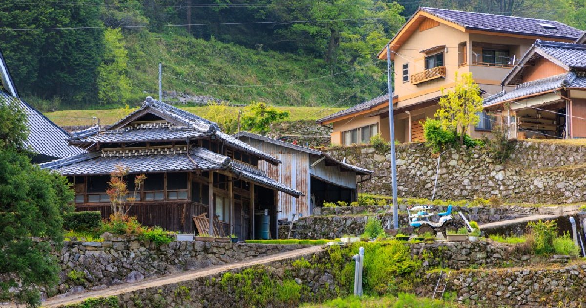 Fotografie a unui sat tradițional japonez cu case rustice din lemn, ziduri de piatră și vegetație luxuriantă.