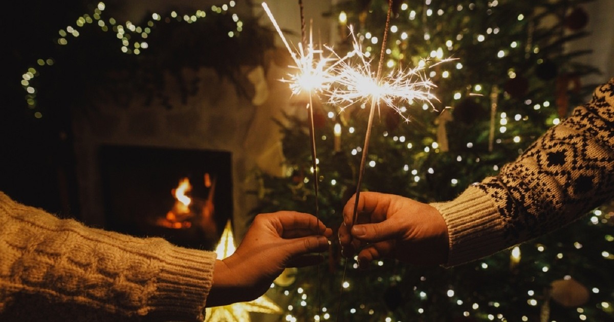 Superstiții de Revelion. Ce să faci în noaptea dintre ani pentru a avea noroc - Prim-plan cu mâini care țin artificii strălucitoare, într-o atmosferă caldă de sărbătoare, cu brad de Crăciun luminat și foc aprins în șemineu