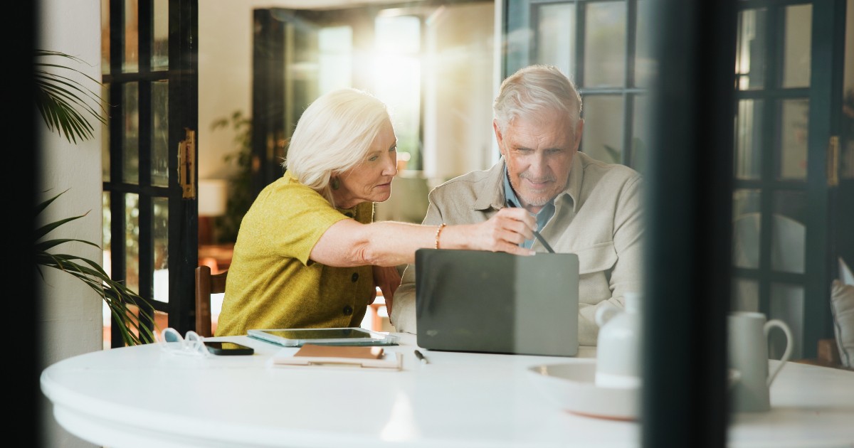 În imagine apare un cuplu de pensionari așezat la masa din bucătărie, analizând împreună informații pe un laptop și documente. Atmosfera sugerează că cei doi își gestionează cu atenție bugetul sau își planifică finanțele, colaborând într-un mod calm și organizat.