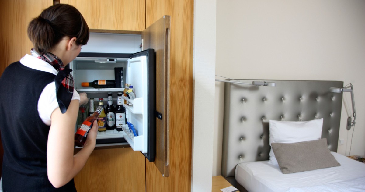 Cameristă de hotel cu uniformă și părul prins care verifică minibarul dintr-o cameră.