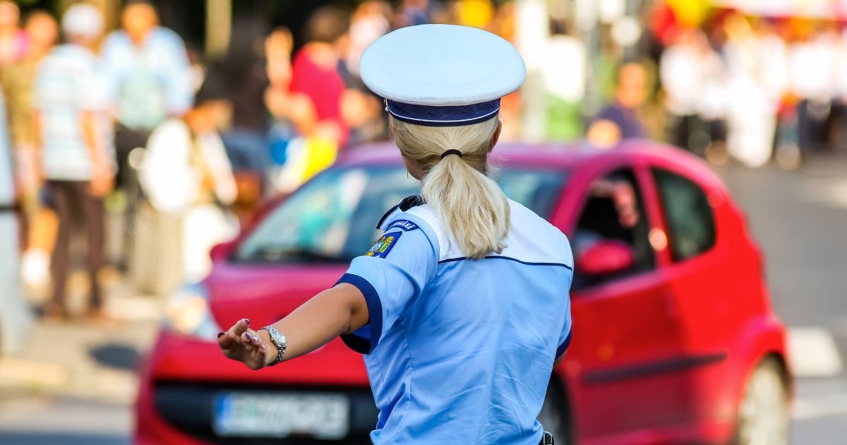 Polițistă îmbrăcată în uniformă de vară, cu o cămașă albastră cu mânecă scurtă, fotografiată din spate, în timp ce dirijează traficul într-o intersecție, având pe fundal o mașină de culoare roșie și oameni pe trotuar