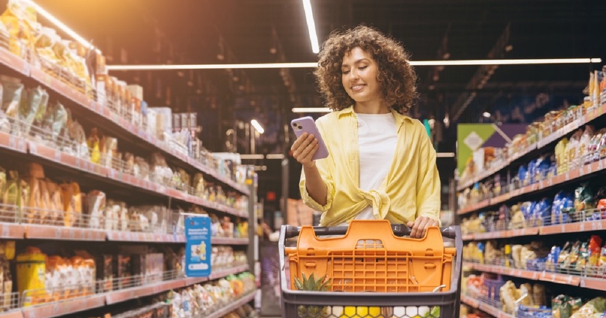 Ghidul cumpărăturilor inteligente: Cum să economisești timp și bani la supermarket - O tânără face cumpărături, împingând un cărucior și verificând lista de cumpărături pe smartphone, pe culoarul unui supermarket