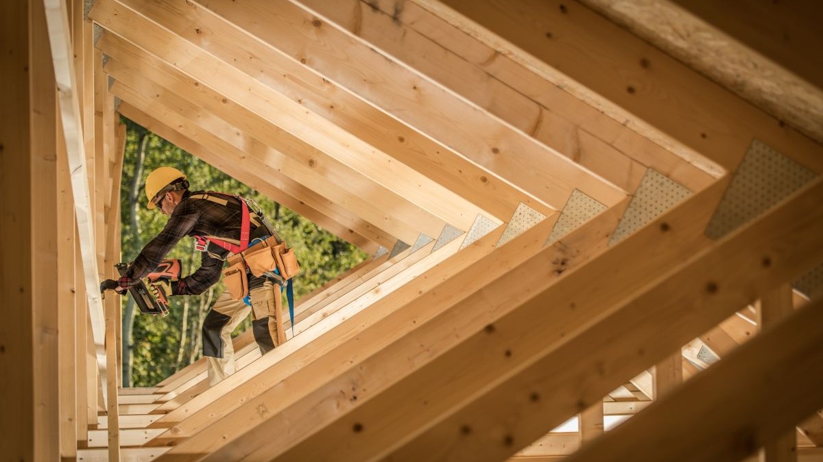 Un muncitor în construcții asamblează cu grijă structura din lemn a unei clădiri, punând în valoare unghiuri și grinzi complexe sub lumina naturală a soarelui, în aer liber.