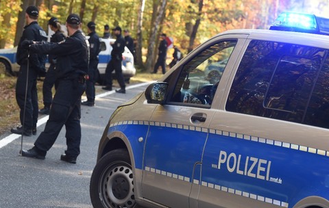 Mai mulți polițiști germani în uniforme desfășurând o operațiune de căutare sau securizare pe un drum de lângă o pădure, având o autospecială de poliție cu girofarul aprins în prim-plan.