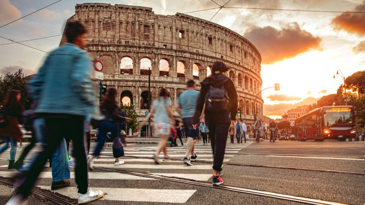 Imaginea prezintă pietoni traversând o stradă pe trecerea de pietoni în fața Colosseumului din Roma, pe fundalul unui apus de soare