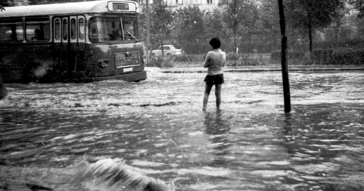 Fotografie alb-negru cu o tânără stâ cu picioarele în apă, pe stradă, în timp ce un autobuz trece și face valuri
