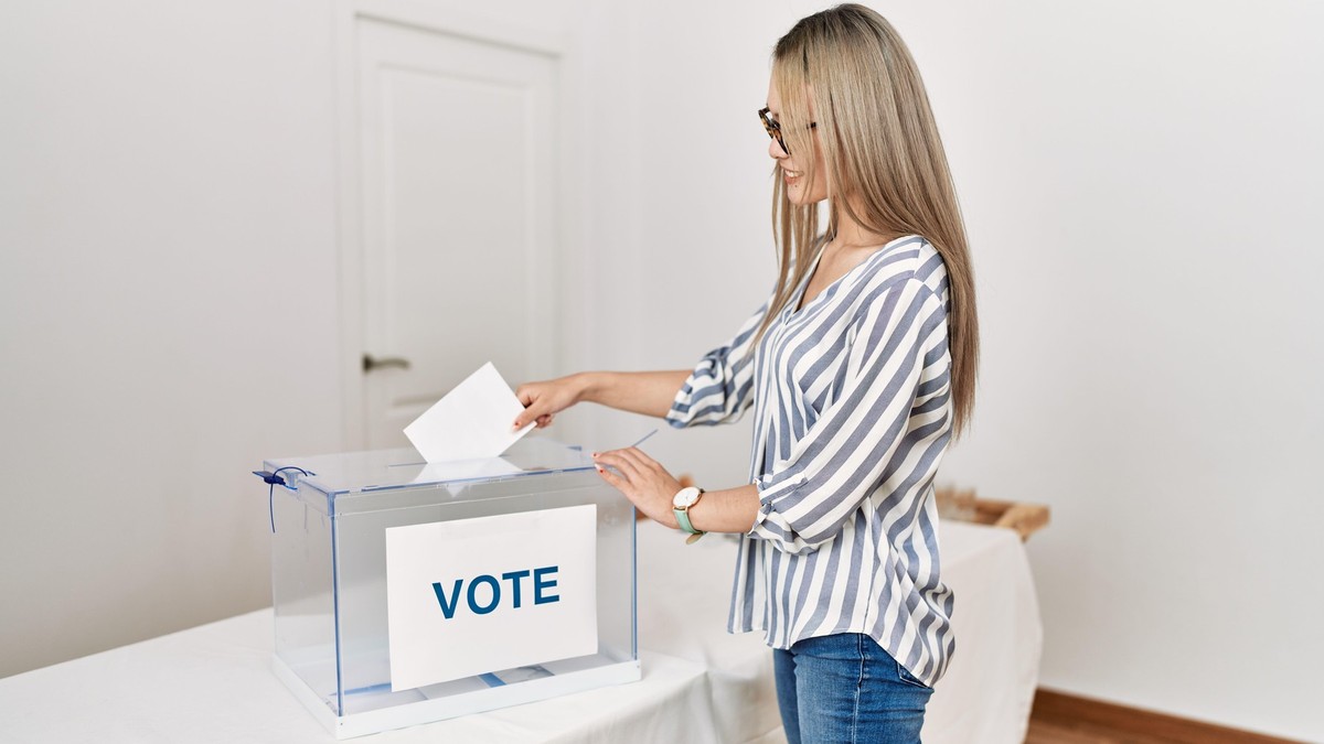 Tânără blondă, care poartă o cămașă albă cu dungi albastre și blugi albaștri, surprinsă în timp ce introduce buletinul de vot într-o urnă